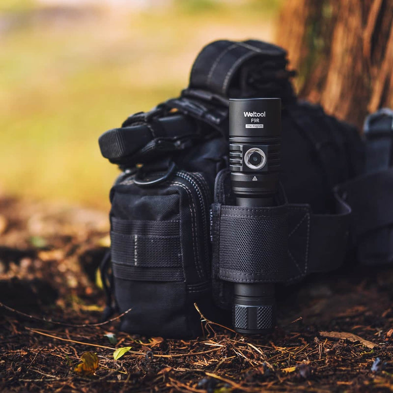 A Weltool F9R high lumen rechargeable LED flashlight is secured in a strap on a black utility bag on the ground near a tree, with blurred outdoor scenery in the background.