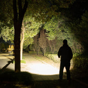 At night, a person stands on a sidewalk illuminated by the powerful Weltool F9R flashlight, with trees and bushes lining the path.