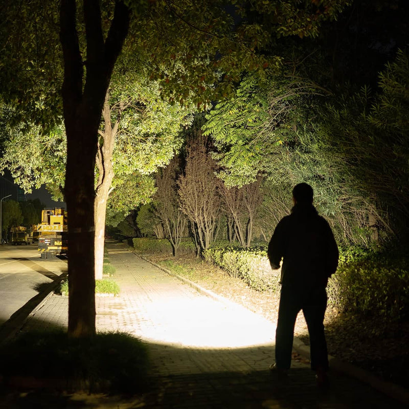 At night, a person stands on a sidewalk illuminated by the powerful Weltool F9R flashlight, with trees and bushes lining the path.