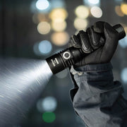 A gloved hand holds and operates the Weltool F9R, a black rechargeable LED flashlight, casting a bright beam in a dimly lit, blurred indoor environment.
