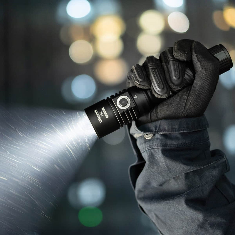 A gloved hand holds and operates the Weltool F9R, a black rechargeable LED flashlight, casting a bright beam in a dimly lit, blurred indoor environment.