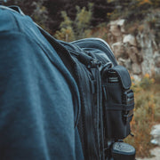 Close-up of a person outdoors near rocks and greenery, wearing a dark blue hoodie and carrying a black backpack with attached gear, including the Weltool FH1 FH2 V2.0 CORDURA Flashlight Holster.