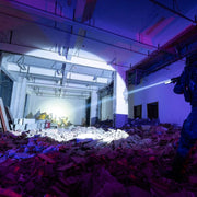A person in tactical gear illuminates rubble and debris inside a damaged building with exposed pipes using a Weltool LH20 weaponlight, known for its high-candela output and aluminum reflector.