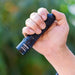A hand holds a black Weltool M7 High-CRI LED flashlight outdoors against a blurred green background. This model offers a 4000K color temperature for natural, accurate lighting.