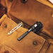 A Weltool M7 High-CRI black LED flashlight with a pocket clip sits in the front pocket of a brown canvas bag beside a metal keychain clip.