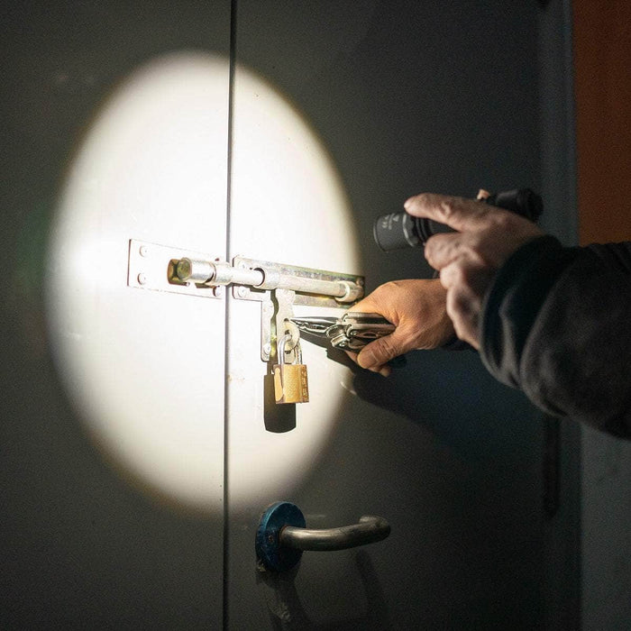 A person holding pliers and a Weltool M7 High-CRI LED flashlight (4000K) attempts to open a locked door secured with a padlock and sliding bolt.