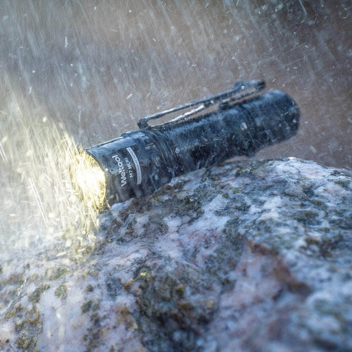 The Weltool M7 High-CRI, a black LED flashlight with high color rendering, shines on a wet rock under bright light as rain falls around it.