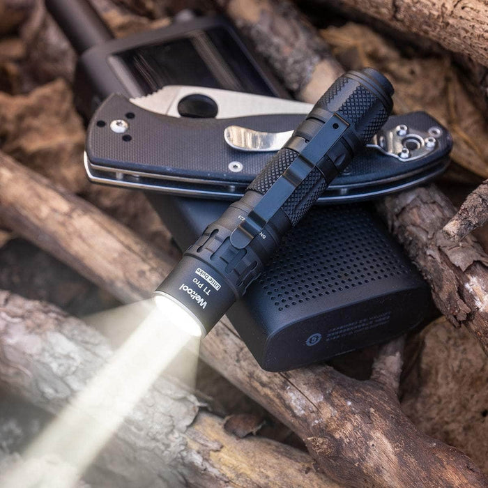 A Weltool T1 Pro TAC illuminating a pile of various outdoor gadgets including a pocket knife, smartphone, and portable speaker on wooden logs.