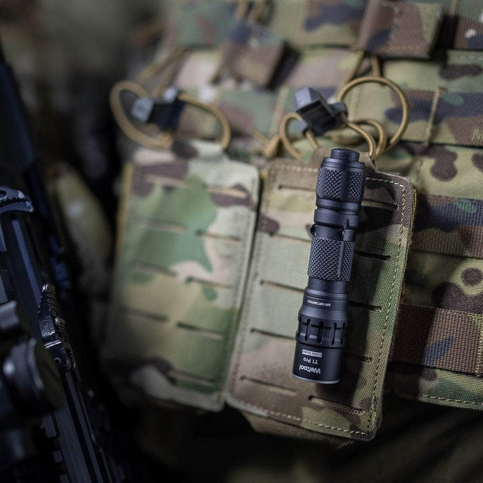 A Weltool T1 Pro TAC flashlight attached to a camouflage military backpack, with blurred parts of a rifle visible on the side.