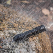 The Weltool T1 Pro V2 flashlight, powered by a 14500 lithium-ion battery, lies on a wet, rocky surface in the rain, perfect for everyday carry.