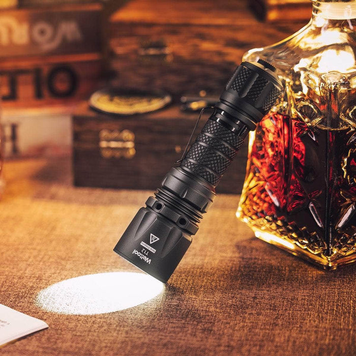 A Weltool T12, emitting a powerful beam of lumens, is positioned on a table alongside a bottle of whiskey.