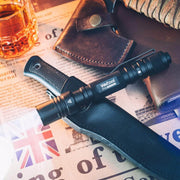 A Weltool T15 pen-style LED tactical flashlight with a rechargeable lithium-ion battery and a sheathed knife rest on a newspaper, next to a leather pouch, a glass of whisky, and a folded cloth.