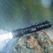 A Weltool T15 LED tactical flashlight emits light while resting on a wet rock, being sprayed with water.