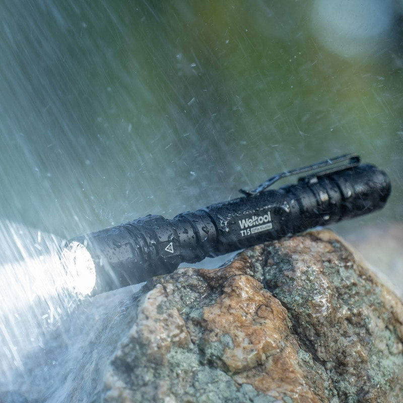 A Weltool T15 LED tactical flashlight emits light while resting on a wet rock, being sprayed with water.