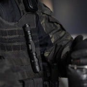 A Weltool T15 rechargeable LED tactical flashlight is clipped to the chest of a person dressed in dark camouflage body armor and tactical gear.
