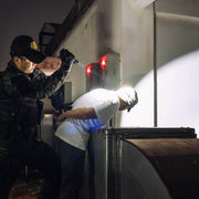 A police officer detains a person against a wall at night, restraining their hands behind their back while illuminating them with a Weltool T15 pen-style flashlight.