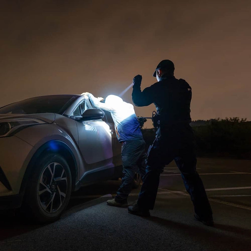 A police officer detains a person against a car at night, illuminating the scene with the high-candela beam of a Weltool T16A compact long-throw LED tactical flashlight. The encounter takes place in a parking lot beneath a cloudy, dark sky.