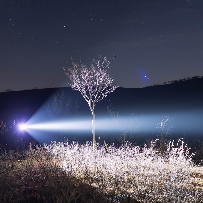 A bare tree stands in a field at night, illuminated by a bright beam of light from the left—perhaps from a Weltool T16B V2 high-lumen flashlight—while hills and stars are visible in the background.
