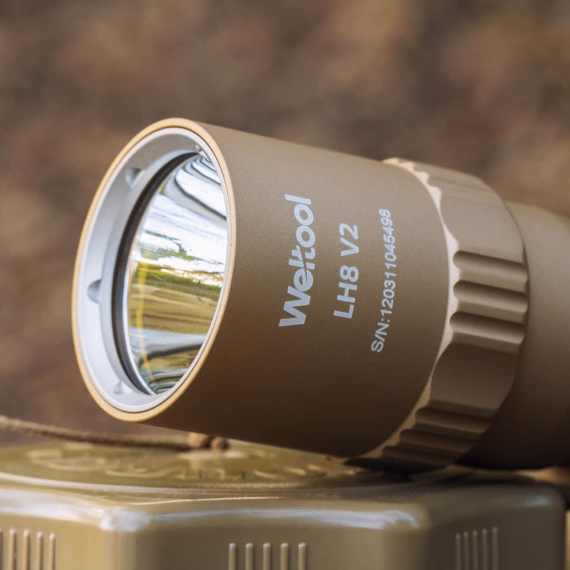 Close-up of a tan Weltool T16B V2 18350 LED flashlight showing the reflective lens and serial number printed on the side, placed on a textured tan surface.