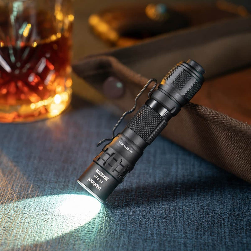 The Weltool T1Pro V3, a compact black flashlight, stands upright on a blue surface shining brightly, with a glass of amber liquid and a brown bag behind—ideal for emergency lighting in any environment.