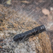 The Weltool T1Pro V3 mini flashlight, powered by a 14500 lithium-ion battery, sits on a wet rock and is sprayed with water—demonstrating its waterproof design for reliable emergency lighting in any condition.