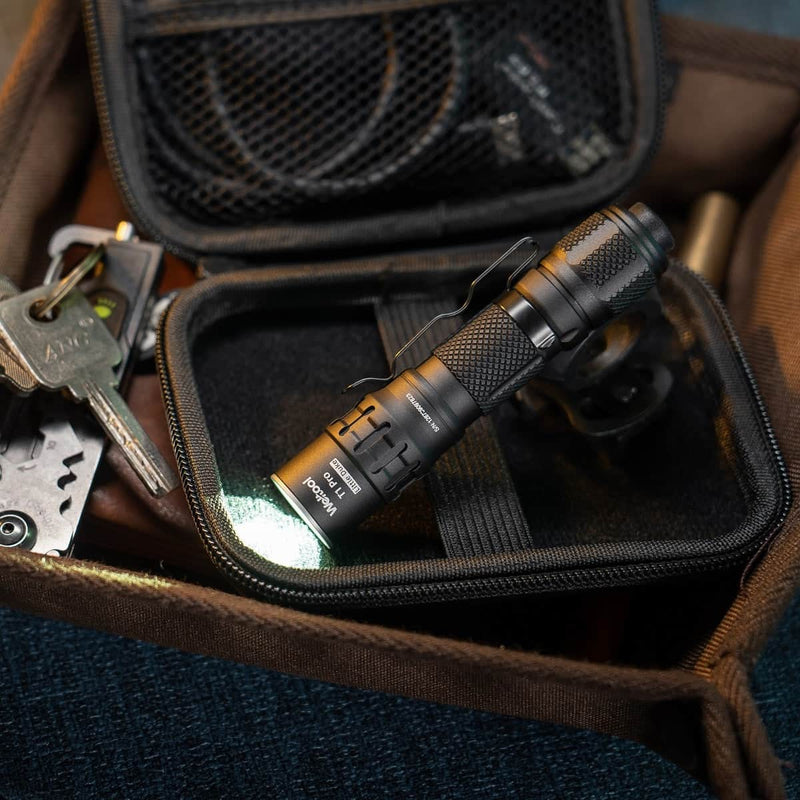 A compact black Weltool T1Pro V3 flashlight, powered by a 14500 lithium-ion battery, is switched on and resting on a zippered pouch next to a set of keys and other everyday items.