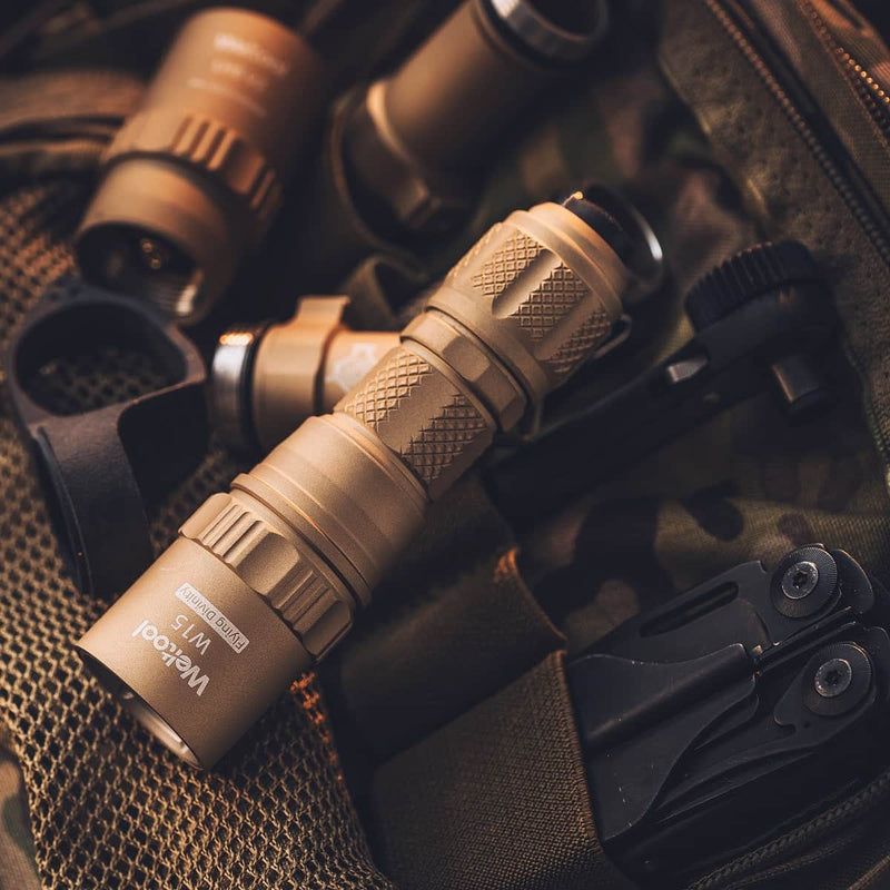 A tan multitool, a Weltool W15 “Flying Divinity” LEP Tactical Flashlight, and other gear are arranged on a camouflage backpack.
