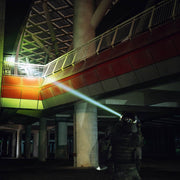 Wearing tactical gear at night, a person aims the Weltool W15 “Flying Divinity” LEP Tactical Flashlight upward, illuminating a multi-level structure with red and white panels.
