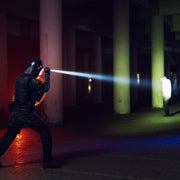 A police officer in tactical gear uses a Weltool W15 “Flying Divinity” LEP Tactical Flashlight, shining its long throw beam towards a standing person in a dimly lit area with concrete pillars.