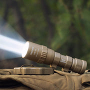The Weltool W35B V2 Compact Weaponlight is turned on, casting a bright beam of light while resting on a khaki bag against a softly blurred green outdoor background.