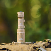 The Weltool W35B V2 Compact Weaponlight in tan stands upright on a khaki bag, with a blurred green outdoor background behind it.