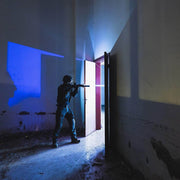 Wearing tactical gear and holding a rifle, a person stands near an open door in a dim, abandoned building lit by the cool glow of the compact Weltool W35S tactical light.