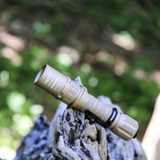 The tan-colored Weltool W3Pro TAC LEP Tactical Flashlight rests on a weathered tree branch outdoors, set against a blurred green background that highlights its sleek, long-range design.