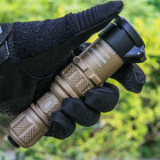 A gloved hand holds a Weltool W3Pro TAC Mini - FDE, a compact tactical flashlight with a textured grip, outdoors with greenery in the background.