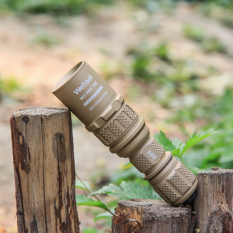 A flat dark earth Weltool W3Pro TAC Mini flashlight is balanced horizontally on two cut wooden posts outdoors, surrounded by green plants and blurred earth in the background.