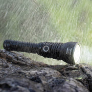 The Weltool W5R Pro LEP rechargeable flashlight sits on a wet rock, glowing brightly as heavy rain falls, showcasing its waterproof design and USB-C charging port.