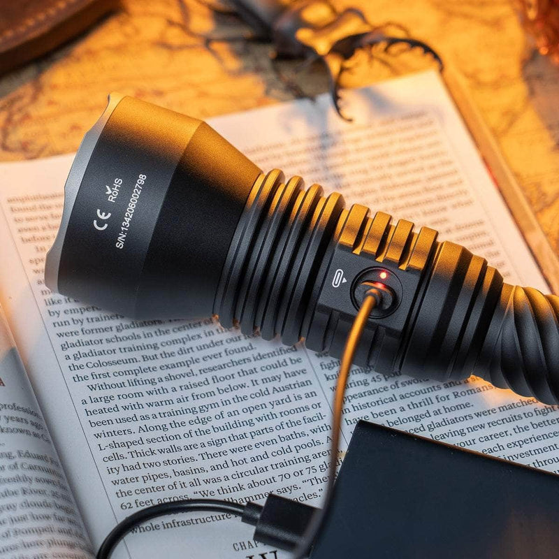 The black Weltool W5R Pro LEP rechargeable flashlight is being charged with a USB-C cable as it rests on an open book with visible text, surrounded by warm lighting.