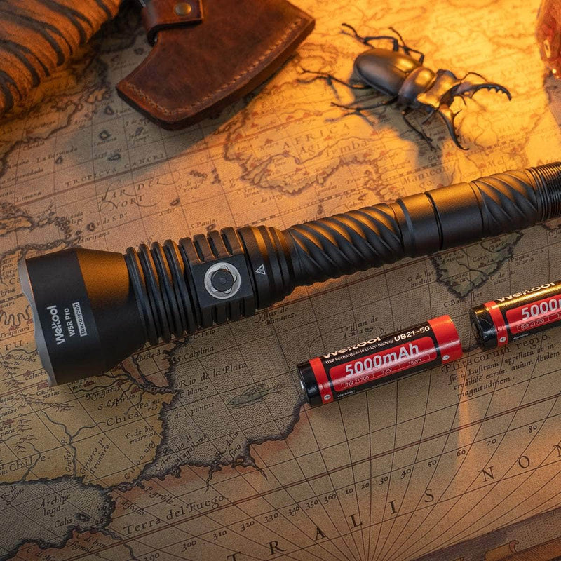 A Weltool W5R Pro LEP rechargeable black flashlight with two red 5000mAh batteries sits on an old world map near a toy beetle and leather pouch. This waterproof USB-C charging flashlight is ideal for adventurers.