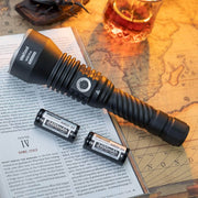 A black Weltool W5R Pro LEP long range flashlight and two 2400mAh rechargeable batteries rest on an open book and a vintage map, with glasses and a drink in the background.