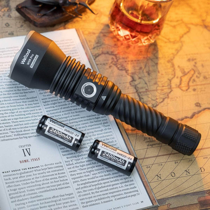 A black Weltool W5R Pro LEP long range flashlight and two 2400mAh rechargeable batteries rest on an open book and a vintage map, with glasses and a drink in the background.