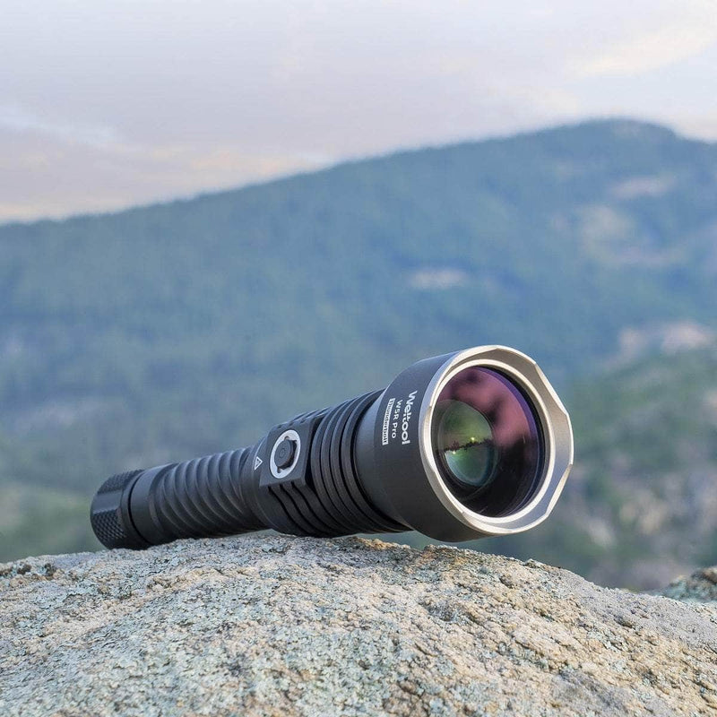 The Weltool W5R Pro LEP black rechargeable flashlight with a large lens sits on a rock outdoors against a blurred green mountain backdrop, showcasing its powerful throw capabilities.