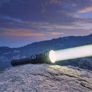 The Weltool W5R Pro LEP emits a bright beam while resting on a rocky surface, with hills and a vibrant dusk sky in the background.