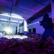 Clad in tactical gear and carrying a rifle, a person uses the Weltool W65S high candela flashlight to navigate a debris-filled, partially destroyed building at night.