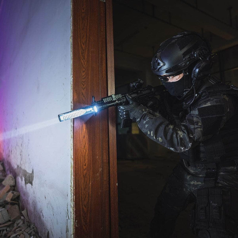 Wearing tactical gear and a helmet, a person aims a rifle fitted with the Weltool W65S high candela flashlight while standing by a doorway in a dim, abandoned building.