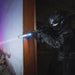 Wearing tactical gear and a helmet, a person aims a rifle fitted with the Weltool W65S high candela flashlight while standing by a doorway in a dim, abandoned building.