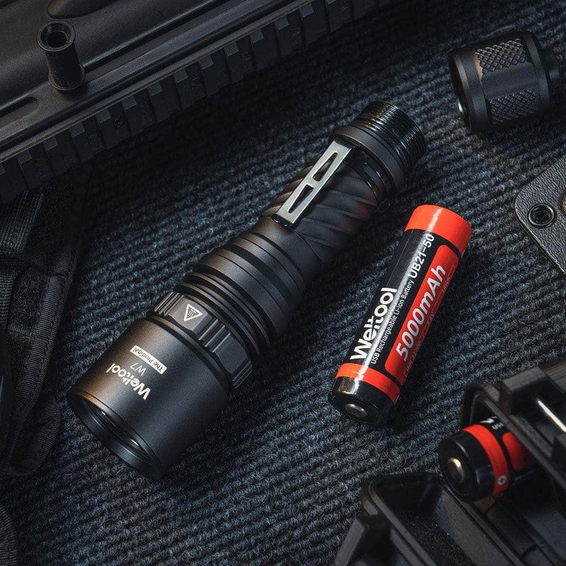 A black Weltool W7 LEP long-range tactical flashlight and a red 5000mAh 21700 lithium battery are on a textured dark surface, surrounded by gear. The flashlight delivers up to 700 lumens.