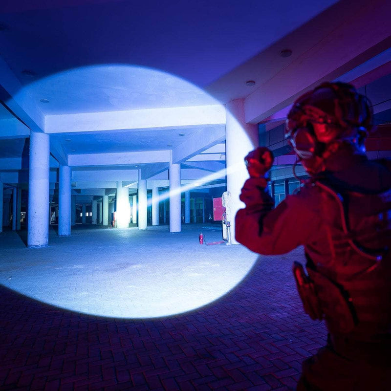 Wearing tactical gear, a person shines the 700-lumen Weltool W7 long-range LEP flashlight into an empty modern building with concrete columns and tiled floors at night.