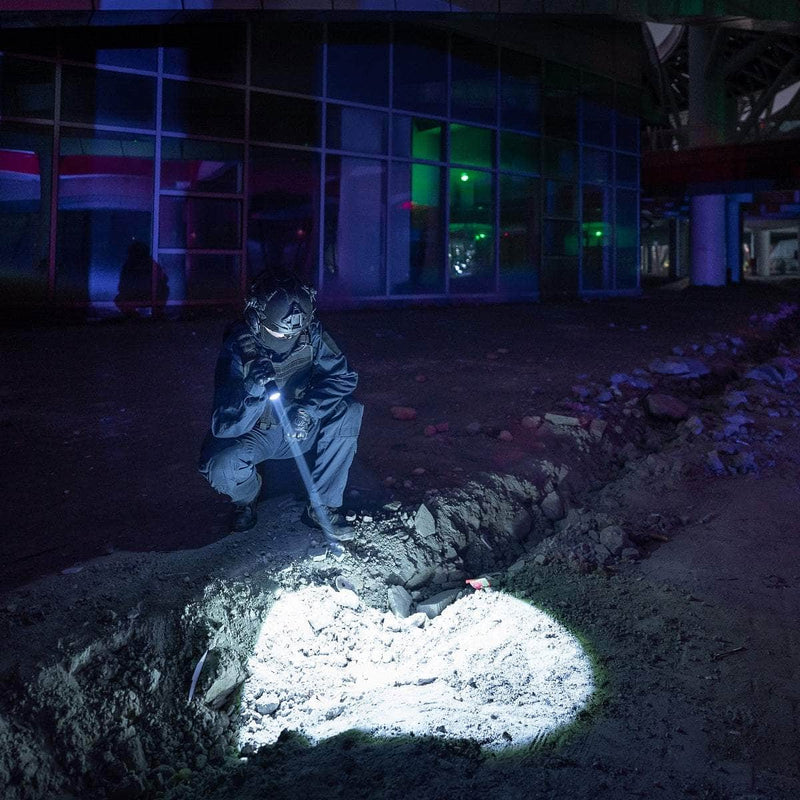 A person in protective gear crouches by a trench at night, inspecting the ground with a Weltool W7 flashlight outside a glass-fronted building.
