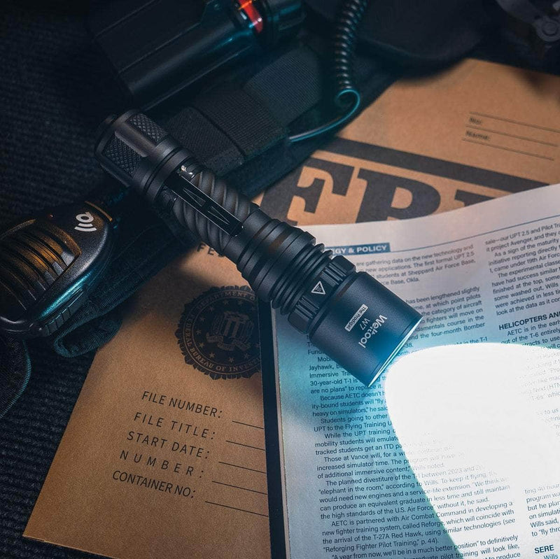 The Weltool W7 LEP tactical flashlight emits 700 lumens, illuminating FBI documents, a radio, and tactical gear on a dark surface for powerful long-range visibility.