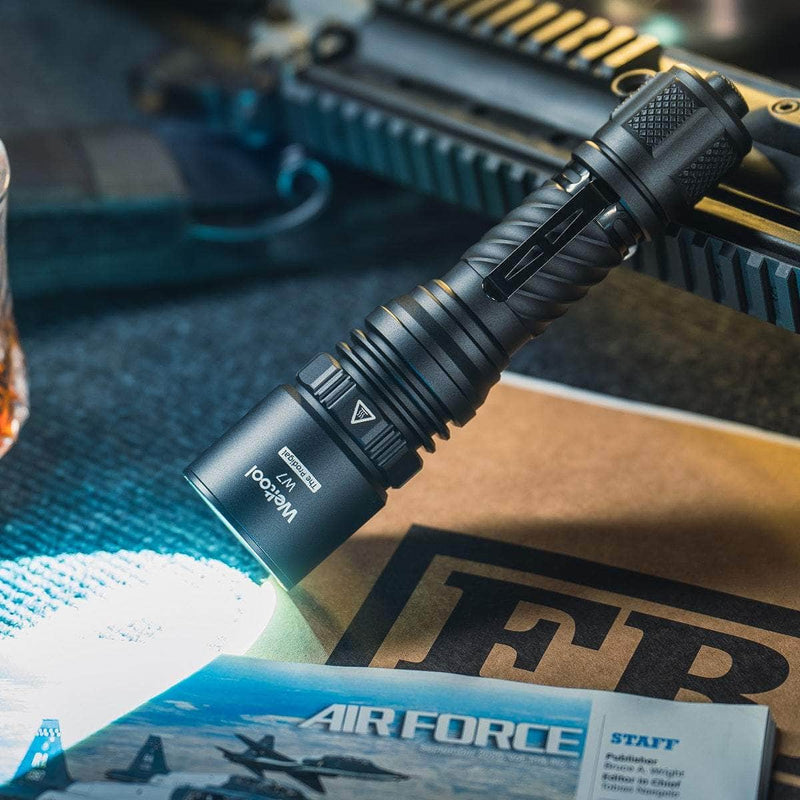 The Weltool W7 long-range tactical flashlight with 700 lumens illuminates papers labeled "FBI" and "AIR FORCE," with part of a firearm seen in the background.
